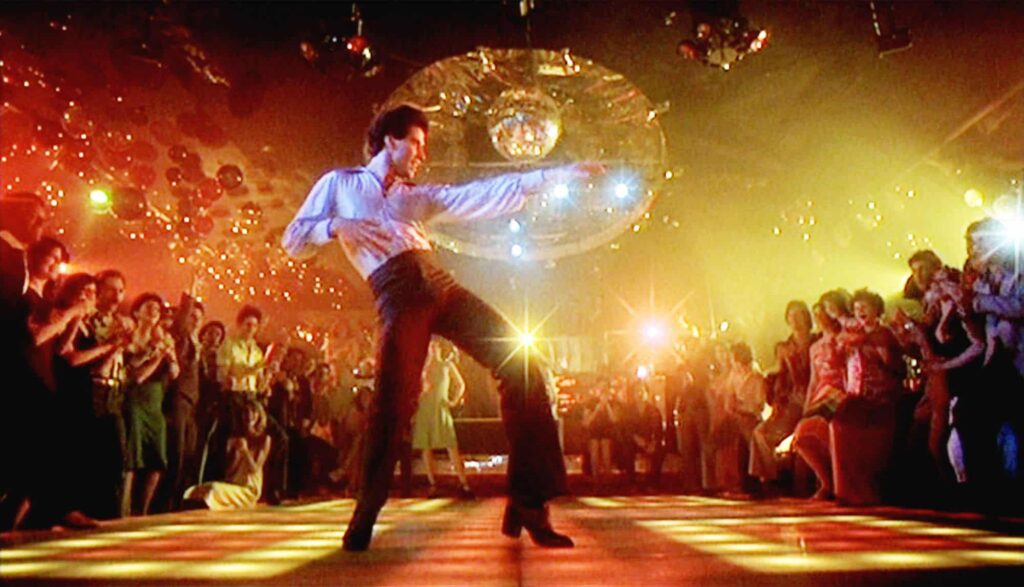 A man in a white shirt and black pants dances energetically on a lit-up dance floor, surrounded by a crowd. A disco ball and colorful lights illuminate the room.