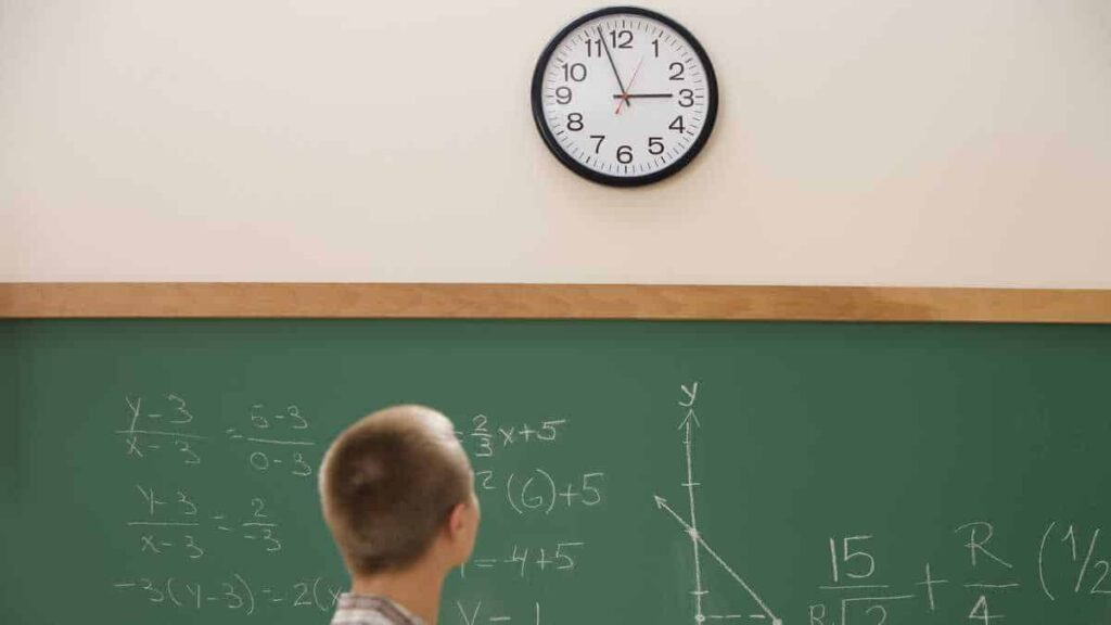 A student looks at a clock on the wall while standing in front of a chalkboard filled with mathematical equations and graphs.
