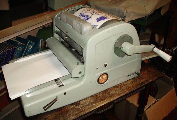 An old-fashioned mimeograph machine with a hand crank, used for duplicating documents, is situated on a wooden table with blank sheets loaded and a stencil attached.