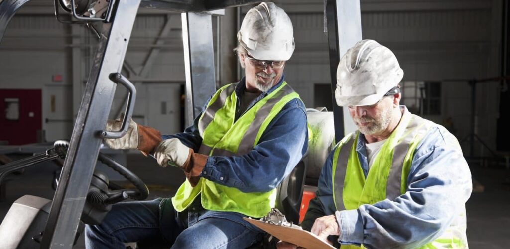 Two warehouse workers wearing safety vests and hard hats review a clipboard while seated on a forklift inside a warehouse.