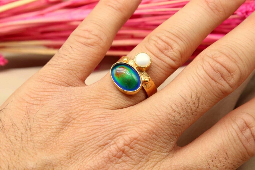 A hand wearing a gold ring with a blue-green oval stone and a smaller white stone, set against a blurred pink background.