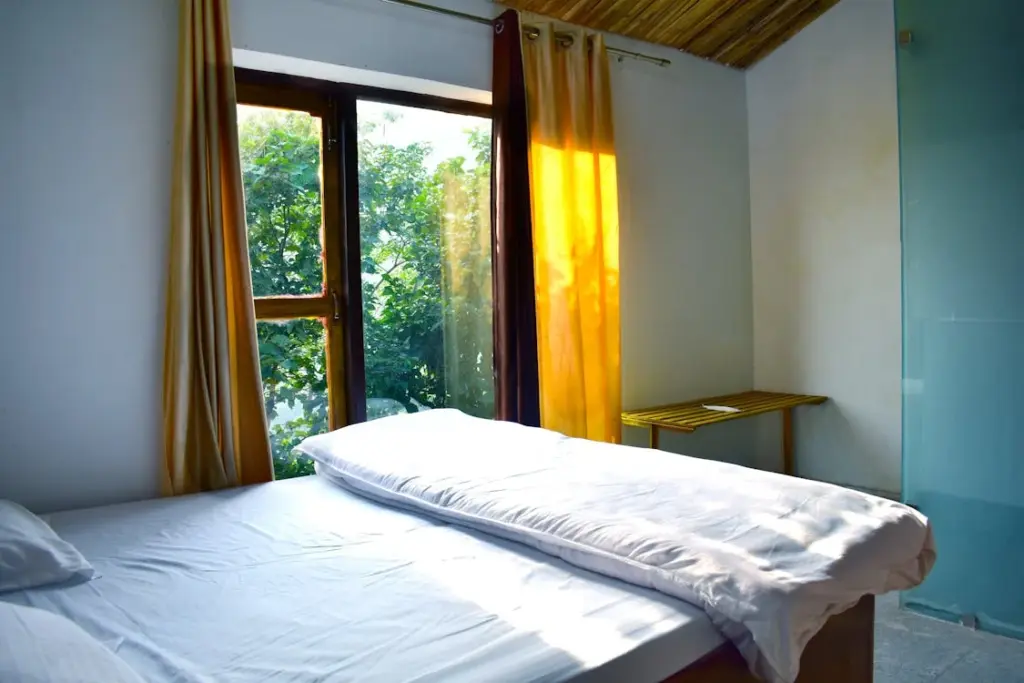 A bedroom with a large window, yellow curtains, a bed with white sheets, and a wooden ceiling. Sunlight streams in through the window, illuminating the room—an effortless way to slash utility bills for busy families.