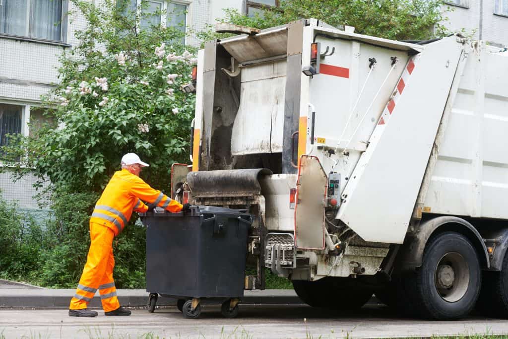 A person in an orange reflective uniform empties a black garbage bin into the back of a white garbage truck on a residential street.
