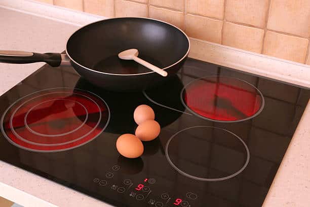 A frying pan with a spatula inside sits on an electric stovetop, with three eggs placed on the stove surface beside it.