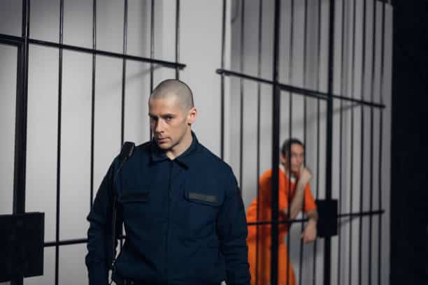 A prison guard in a dark uniform stands outside a cell, while an inmate in an orange jumpsuit sits inside the cell in the background.
