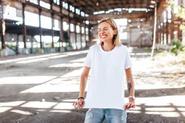 A person wearing a white t-shirt and jeans stands in an abandoned warehouse, smiling brightly.
