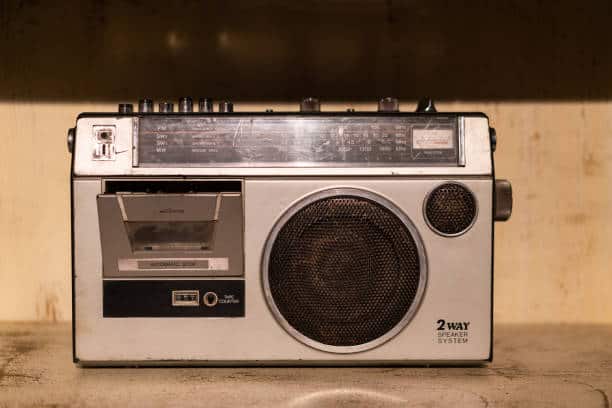 A vintage portable radio with a cassette player and a large speaker on the right side, featuring a worn exterior and various control knobs on the top.