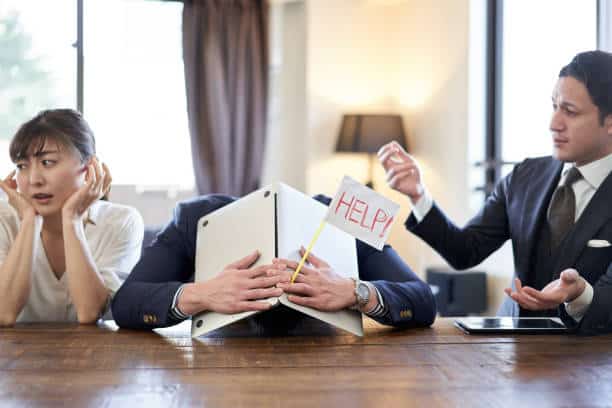 Three people in a meeting room: one looking concerned, one with head on the table holding a "HELP!" sign, and one gesturing while speaking.