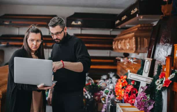 Two people look at a laptop screen in a room filled with coffins and floral arrangements.