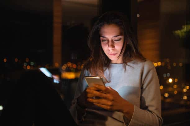 A woman is intently looking at her smartphone screen at night, with city lights blurred in the background.