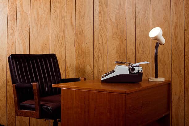 A wooden desk with a typewriter, a small lamp, and a brown leather chair in a wood-paneled room.