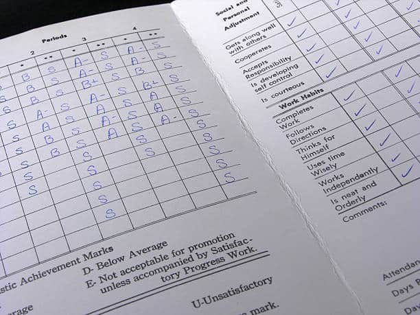 A close-up of a report card showing various academic achievement marks and work habits, including a section with checkmarks for attitudes and effort.