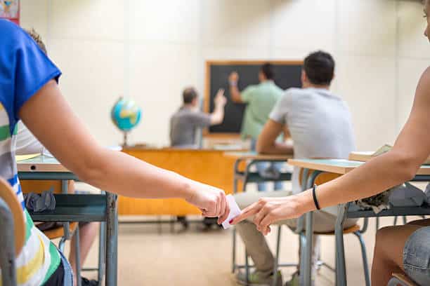 Two students pass a note to each other in a classroom while a teacher writes on a blackboard. Other students are visible sitting at their desks, facing the front of the room.