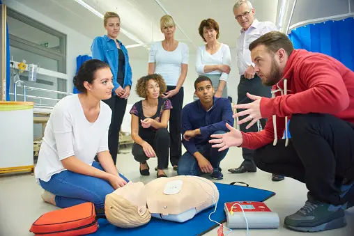 A group of people attends a CPR training session. An instructor in a red hoodie demonstrates the technique using a CPR manikin while participants watch attentively. CPR equipment is visible on the floor.