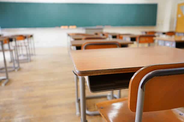 An empty classroom with wooden desks and chairs arranged in rows, facing a green chalkboard. The room has a wooden floor and a bright atmosphere.
