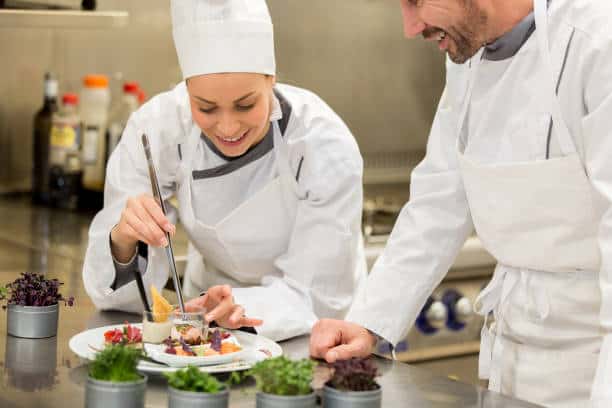 Two chefs in white uniforms and hats are plating a dish in a professional kitchen, surrounded by small containers of herbs and garnishes.