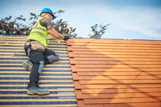 A construction worker wearing a blue helmet and high-visibility vest is installing orange roof tiles on a sloped roof.