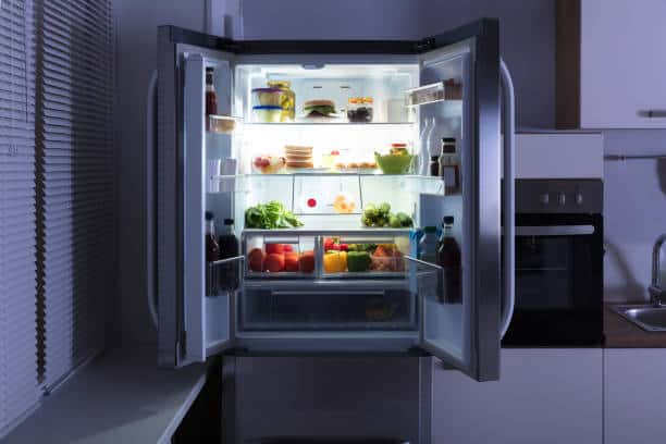 An open refrigerator filled with various types of food, including fruits, vegetables, jars, and bottles, in a modern kitchen at night.
