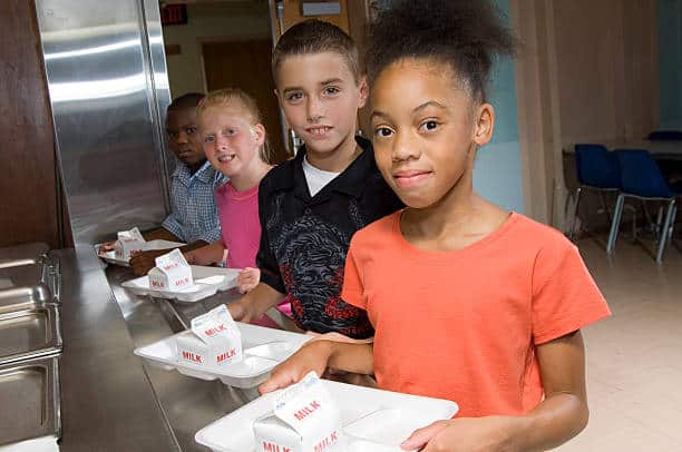 Four children stand in line with lunch trays containing milk cartons in a cafeteria.