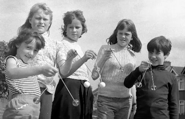 Five children stand outdoors, holding and playing with clackers, a popular toy from the 1970s.