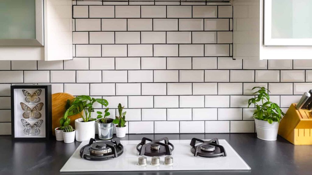 A modern kitchen with a white-tiled backsplash. The countertop features a gas stovetop, small potted plants, a framed butterfly display, and a knife block. White cabinets are overhead.
