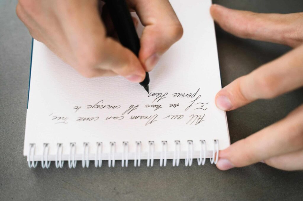 A person is writing calligraphy on a spiral-bound notebook with black ink.