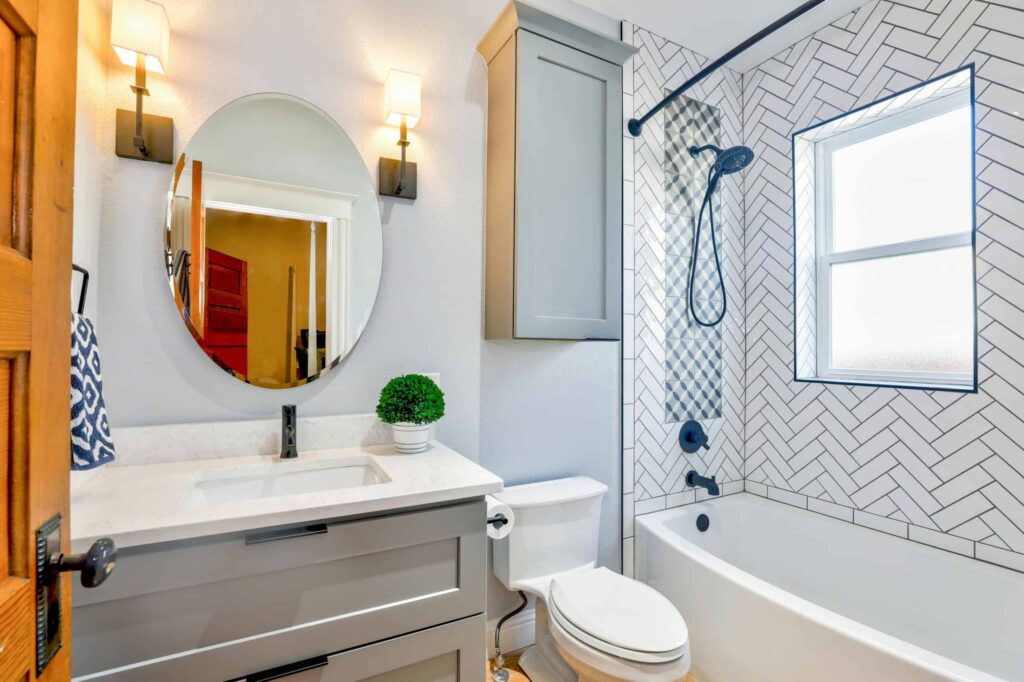 Modern bathroom with a gray vanity, white countertop, oval mirror, wall-mounted lights, toilet, and a bathtub with a herringbone-tiled wall and window.