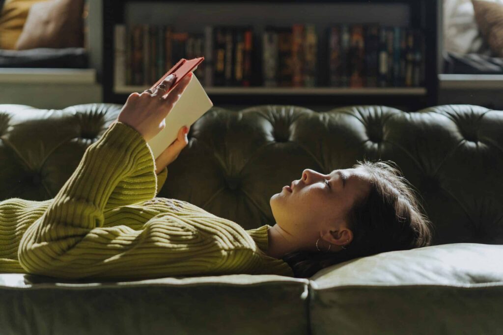 A person lies on a green sofa holding an open book close to their face, with a bookshelf in the background.