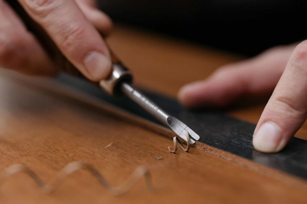 Close-up of hands using a small chisel to carve a curly wood shaving from a brown wooden surface, with a black straightedge used as a guide.