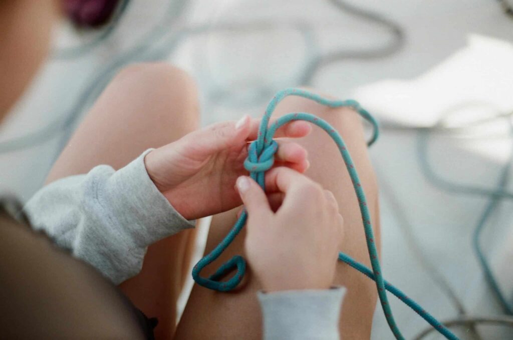 Close-up of a person's hands tying a blue rope in a knot. The individual is wearing a light grey long-sleeve shirt, and their legs are visible in the background.