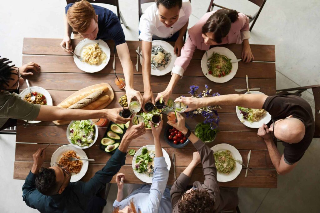Eight people sit around a table sharing a meal, raising their glasses for a toast. Various plates of food, including salads, pasta, bread, and avocados, are spread across the wooden table.