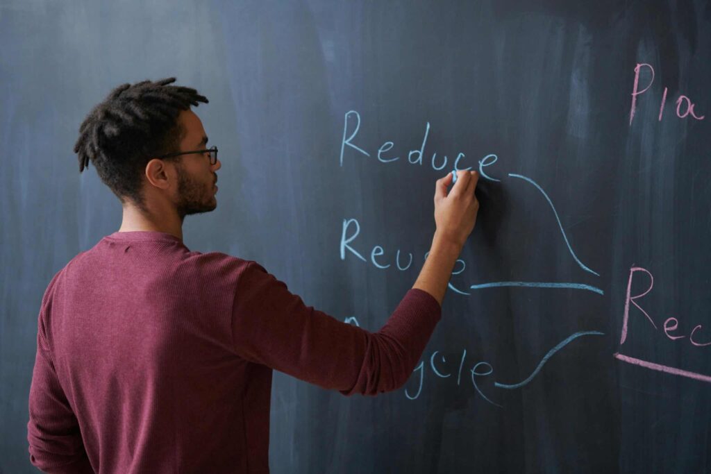 A person writes the words "Reduce," "Reuse," and "Recycle" on a blackboard with blue and pink chalk.
