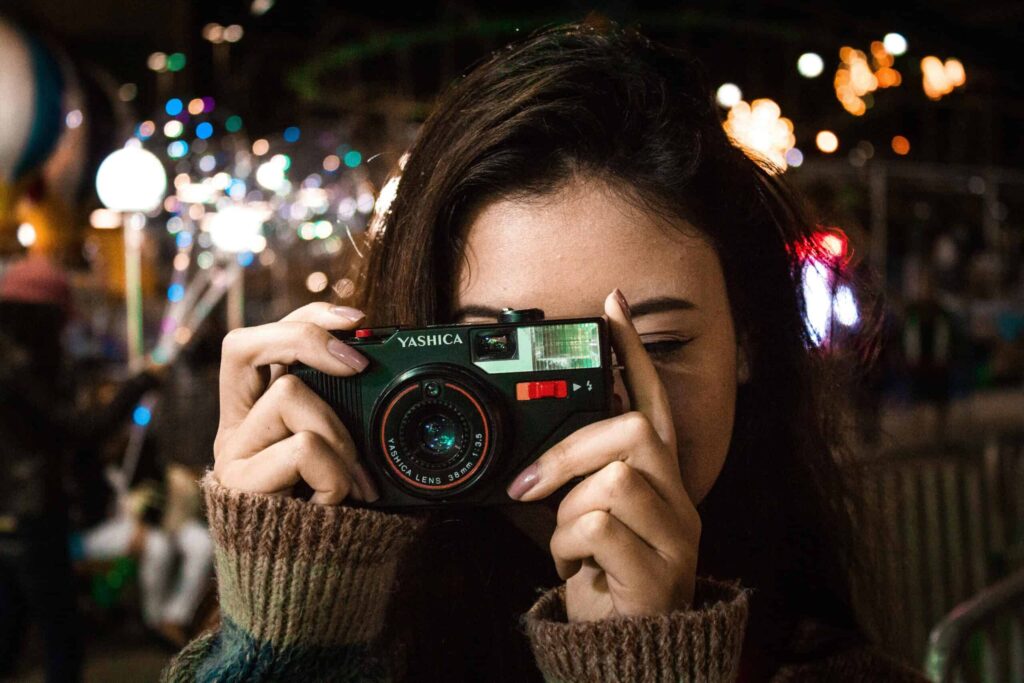 A person wearing a brown sweater holds a Yashica camera up to their face, ready to take a photo. Blurry lights can be seen in the background.