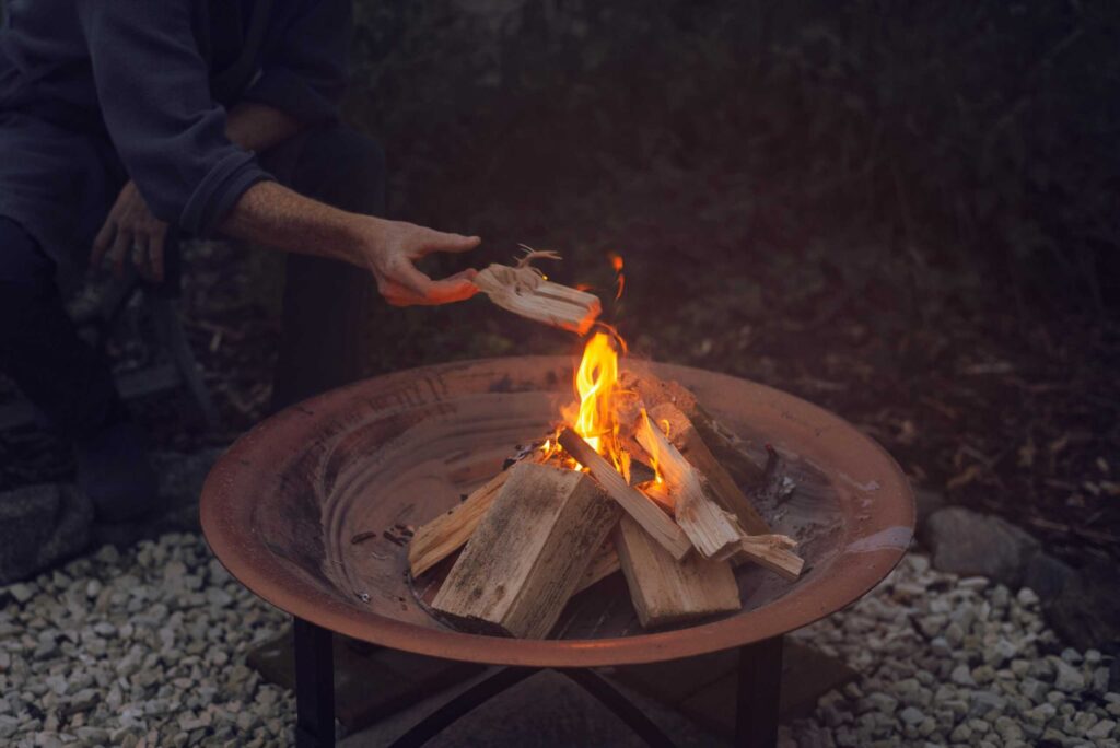 A person is adding a piece of wood to a burning fire in a round metal fire pit.
