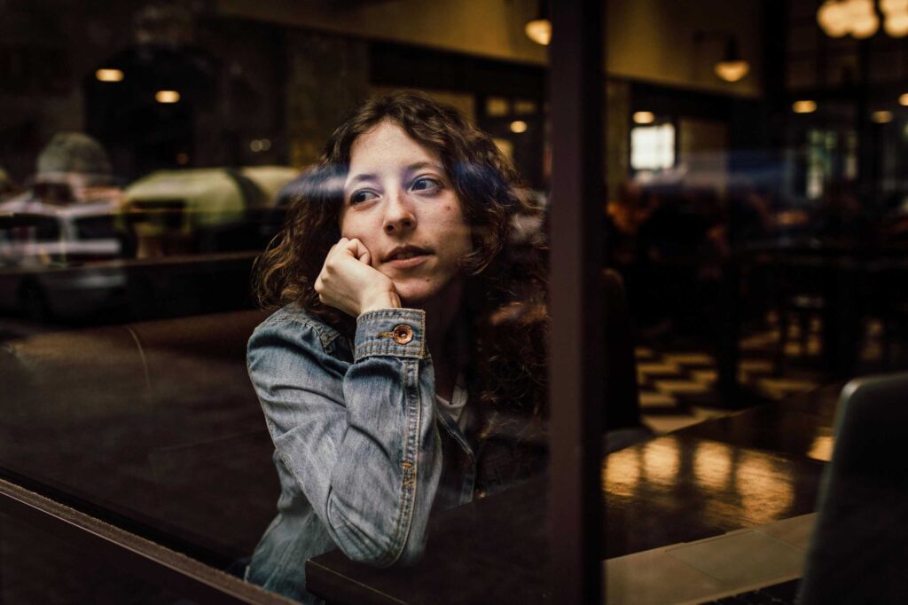 A woman with curly hair looks out of a cafe window, resting her head on her hand, appearing thoughtful. She is seated at a table wearing a denim jacket.