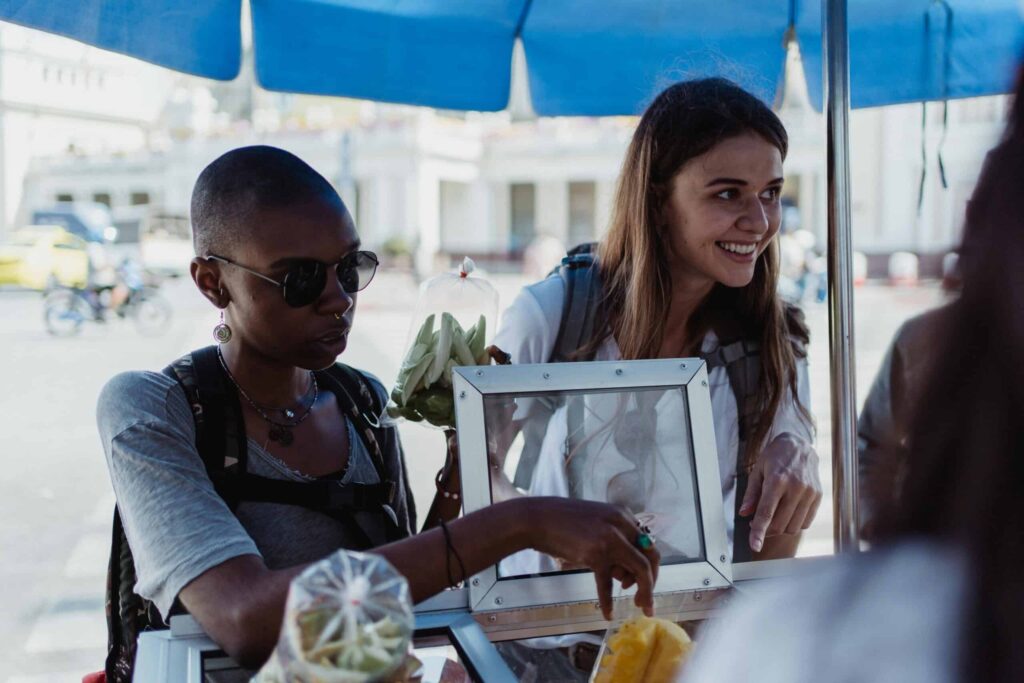 Two people stand at an outdoor vendor's stall. One is holding a bag and pointing, while the other smiles. An assortment of fresh produce is visible on the counter.