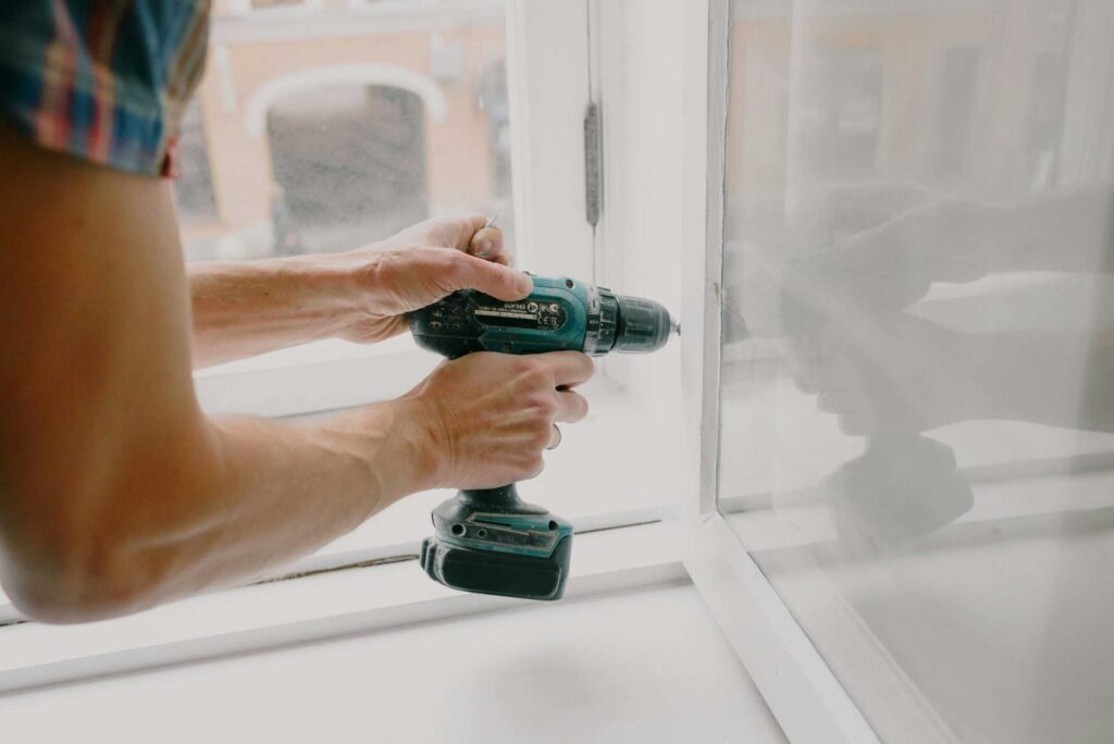 A person using a cordless drill to attach a fixture near a window.