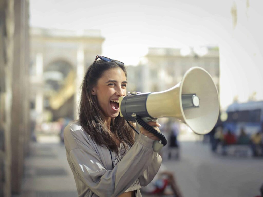 A woman is shouting into a megaphone while standing outside in a city area. She has long dark hair and is wearing a light jacket.