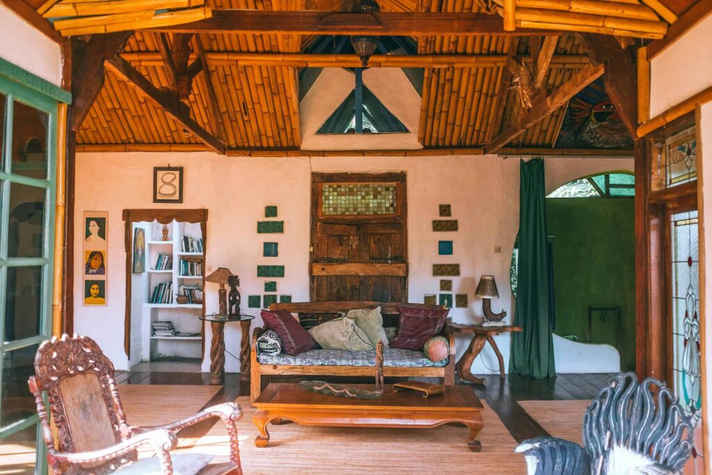 A cozy living room with rustic wooden furniture, a couch, bookshelves, portraits on a wall, a lamp on a side table, and a green curtain partially covering the doorway. The ceiling is made of bamboo.