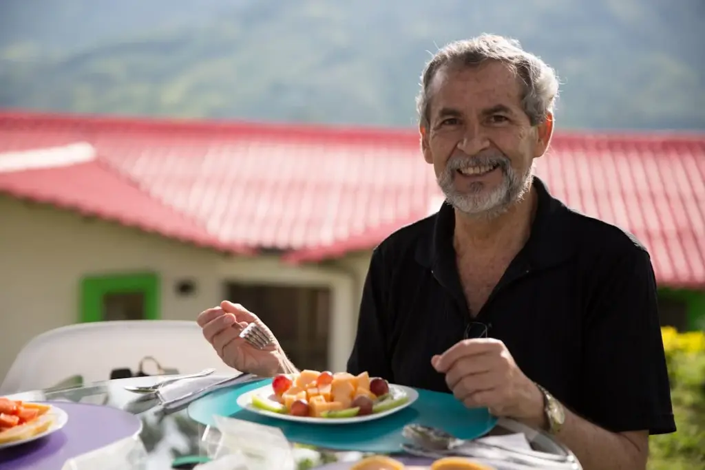 An elderly man with gray hair and beard smiles while holding a fork and sitting at a table with a plate of fruit salad outdoors near a red-roofed building.