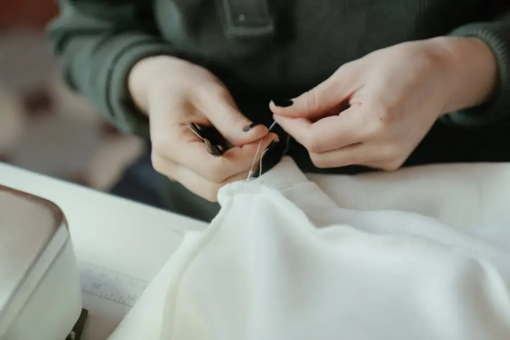 A person wearing a dark long-sleeved shirt is sewing white fabric with a needle and thread.