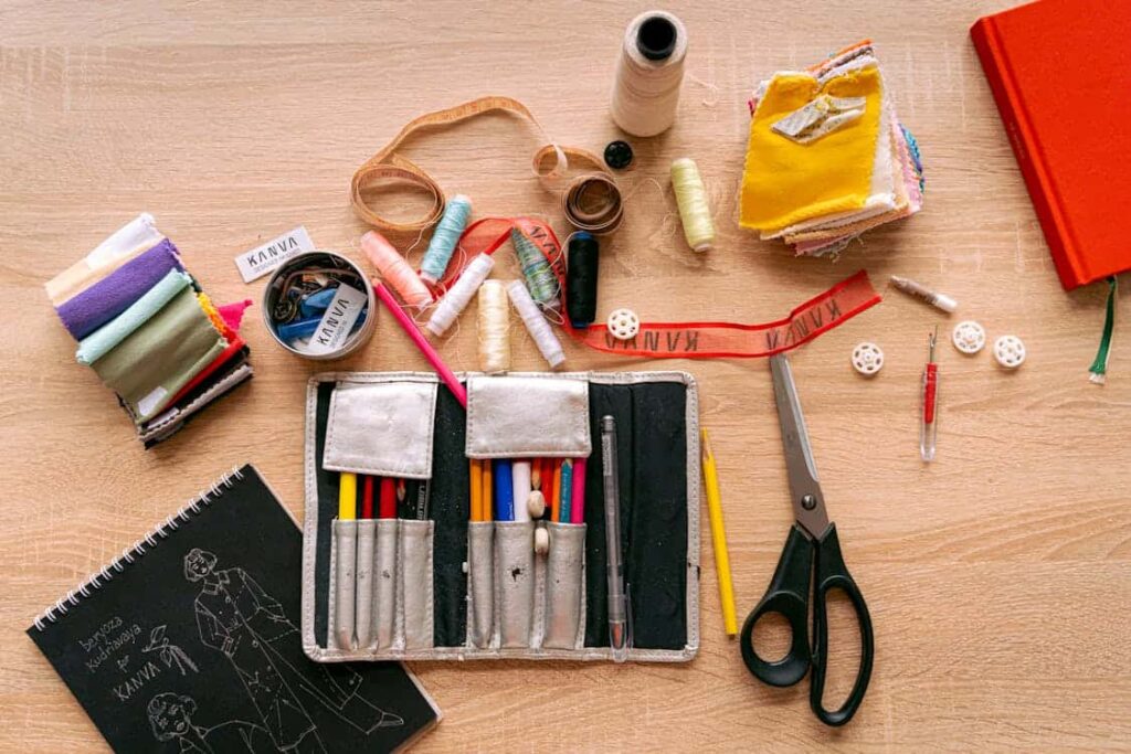 A sewing kit on a wooden surface with thread spools, fabric pieces, scissors, a notebook, colored pencils, ribbon, and various sewing tools.