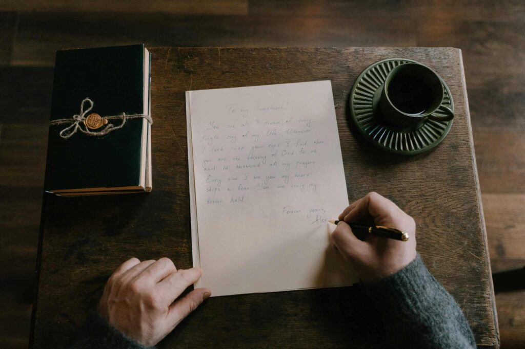 A person writes a handwritten letter on a wooden desk with a closed book tied with string and a cup of coffee beside them.