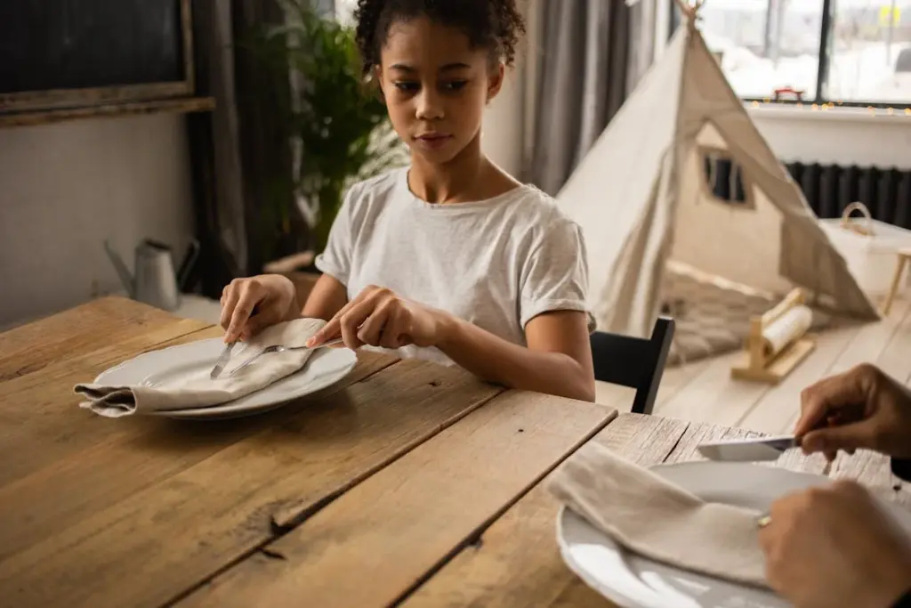 A young person, seated at a table, is unfolding a napkin and arranging silverware on a plate. Another person’s hands are visible, also preparing the table. A fabric teepee is seen in the background.