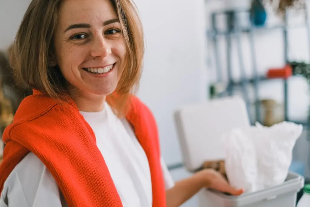A woman with brown hair smiles while holding open a trash bin in a domestic setting. She is wearing a white top with a red sweater draped over her shoulders.