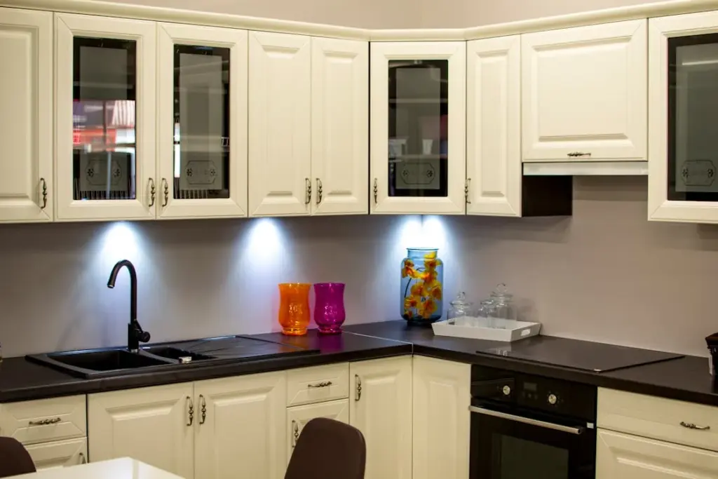 Modern kitchen with white cabinetry, black countertops, and under-cabinet lighting. Orange and pink vases, along with a jar of decor, are placed on the counter near the sink.