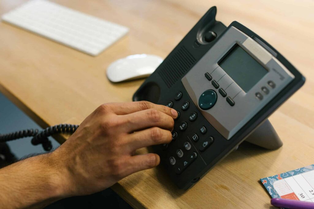 A person dials a number on a desk phone beside a keyboard and mouse on a wooden desk.