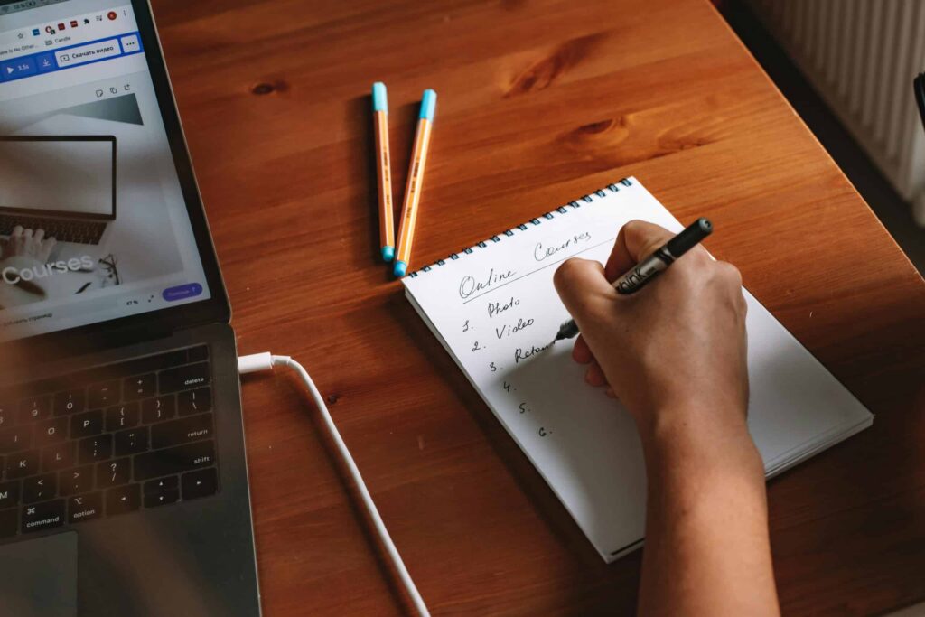 A person writes a list titled "Online Courses" in a notebook next to an open laptop and three pens on a wooden desk.