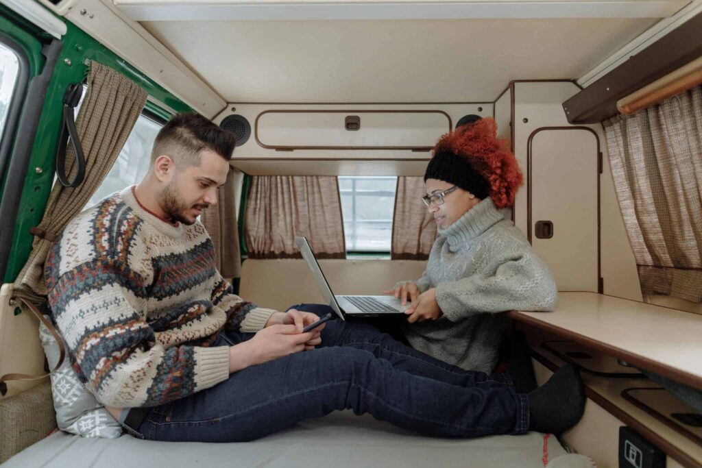 Two people are sitting in a camper van; one uses a smartphone while the other types on a laptop. Both are dressed warmly in sweaters and beanies.