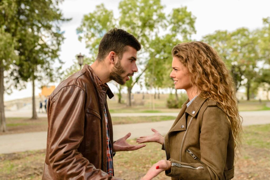 A man and a woman are standing outside in a park, engaged in an intense conversation, both gesturing with their hands. Trees and a path are visible in the background.